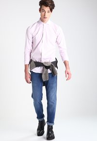 Camisa de botones rosa claro, jeans oscuros y botas ankle negras. Un suéter gris tejido está atado a la cintura. Tejido suave, estilo casual.