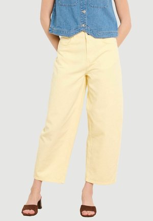 Mujer que lleva pantalones anchos de color amarillo pálido, sandalias de tacón marrones y un chaleco corto de mezclilla azul frente a un fondo liso.