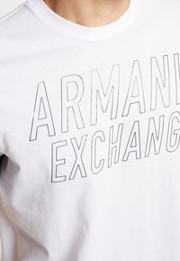 Bílý bavlněný tričko s výrazným černým konturovým potiskem "ARMANI EXCHANGE" na přední straně. Jednoduchý design s kulatým výstřihem.