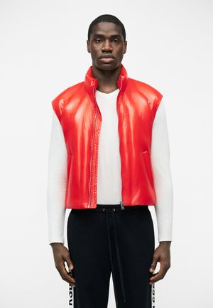 Férfi élénkvörös ujjatlan steppelt mellényben, fehér hosszú ujjú inggel és fekete, oldalt feliratos nadrággal, fehér háttér előtt áll.