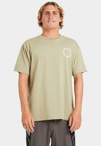 Camiseta de manga corta color verde oliva, hecha de algodón, con un cuello redondo y un pequeño estampado gráfico blanco en el lado izquierdo del frente.