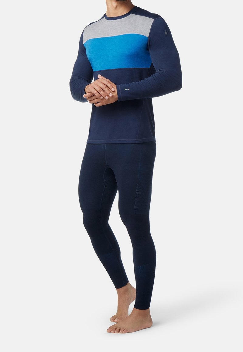 Man i marinblå, blå och grå långärmad tröja samt marinblå tajta leggings, stående barfota mot en enkel vit bakgrund.