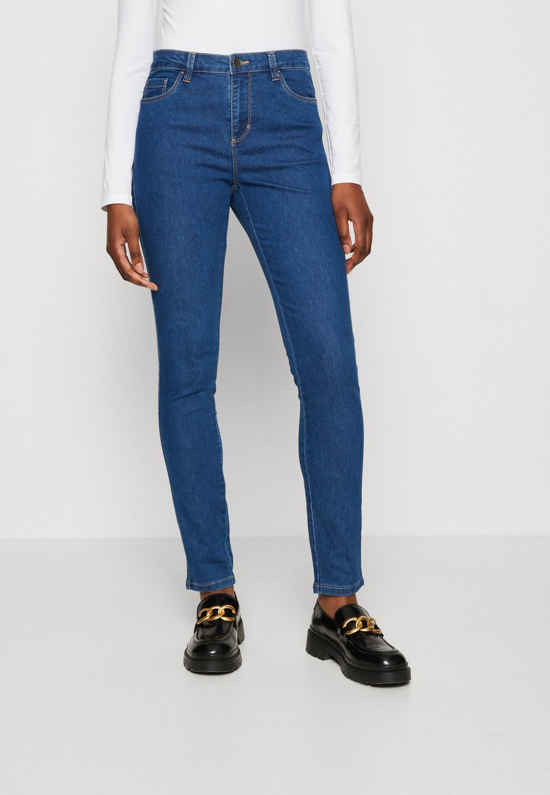 Jeans ajustados de tiro alto en color azul, con cinco bolsillos, costuras en contraste y corte slim, combinados con zapatos negros que tienen un detalle de cadena dorada.