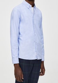 Hellblaues Hemd mit langen Ärmeln, spitzem Kragen und leichter Textur. Kombiniert mit dunkel karierten Hosen, die einen lässigen Stil zeigen.