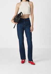 Modré džíny, béžový zkrácený halter top, černá strukturovaná kabelka a červené ploché boty, stojící na jednobarevném pozadí.