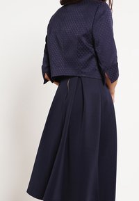 Abito blu navy con giacca cropped a trama; presenta maniche a tre quarti e una gonna plissè. Tessuto liscio con una leggera lucentezza.
