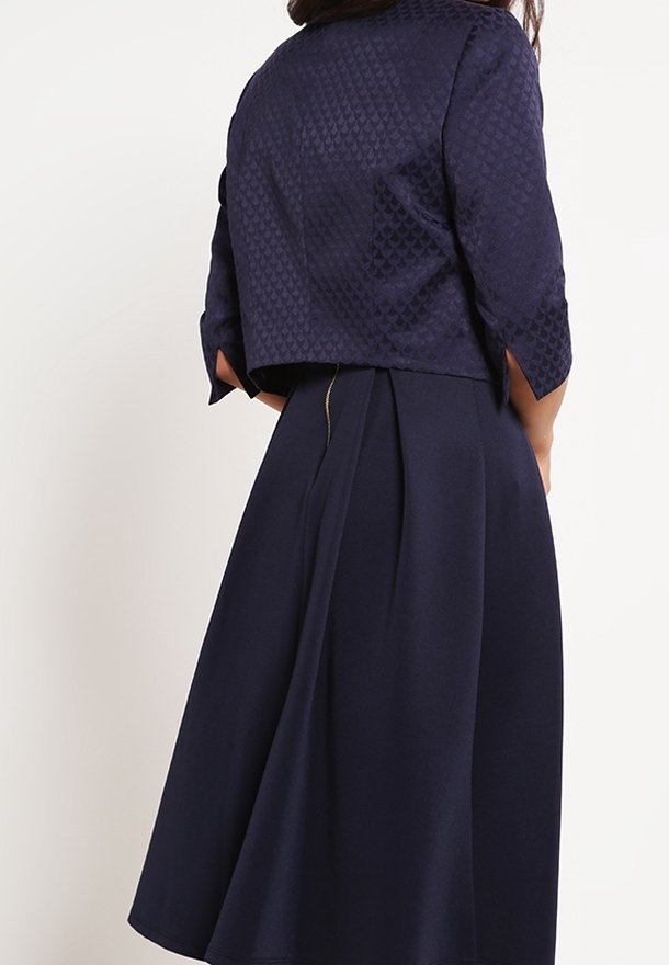 Abito blu navy con giacca cropped a trama; presenta maniche a tre quarti e una gonna plissè. Tessuto liscio con una leggera lucentezza.