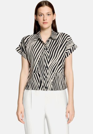Femme portant une chemise boutonnée à manches courtes à rayures zèbre noires et blanches accompagnée d'un pantalon blanc, debout devant un fond uni.
