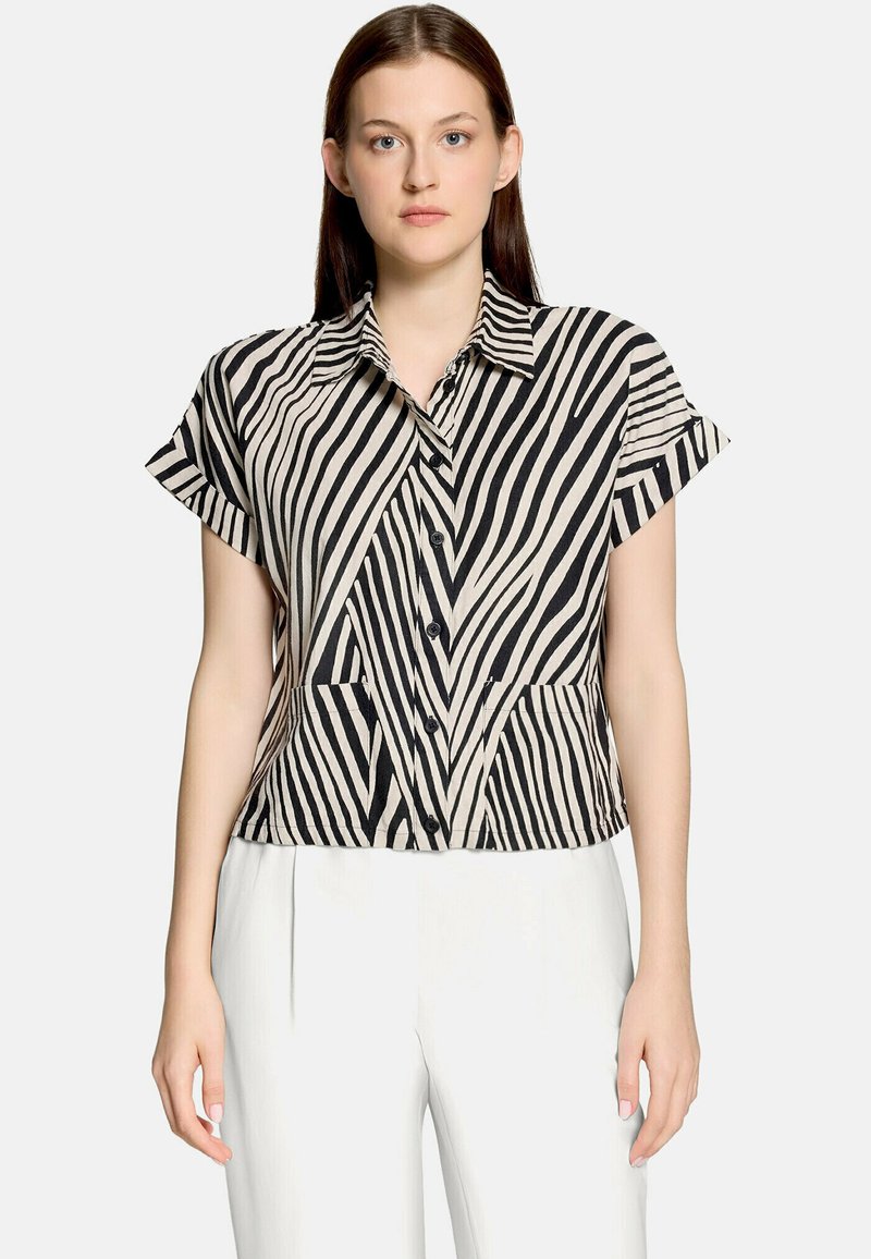 Femme portant une chemise boutonnée à manches courtes à rayures zèbre noires et blanches accompagnée d'un pantalon blanc, debout devant un fond uni.