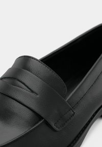 Mocassin en cuir noir avec un détail de bride cousue sur le dessus, texture lisse et bout arrondi.