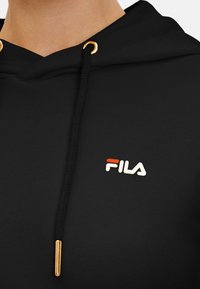 Felpa nera con hardware in accentuazione oro, tessuto strutturato e logo FILA in rosso e bianco posizionato sul davanti.