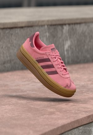 Baskets Adidas Gazelle roses avec des rayures marron et une semelle en gomme flottant au-dessus de marches en béton.