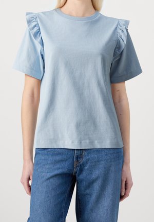 Haut bleu clair à manches courtes avec épaules à volants porté avec un jean bleu, présenté de la taille au cou sur une personne debout.