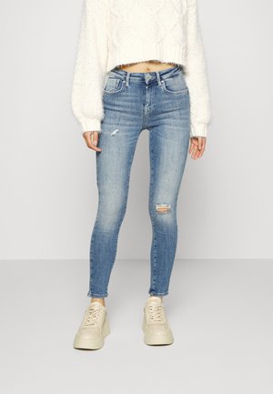 Jean skinny en denim bleu clair avec un genou déchiré, associé à un pull moelleux blanc et des baskets crème, créant un look décontracté.