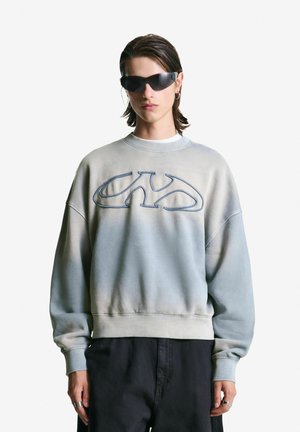 Person wearing oversized gray sweatshirt with abstract logo, black pants, dark sunglasses, and silver rings against a plain white background.