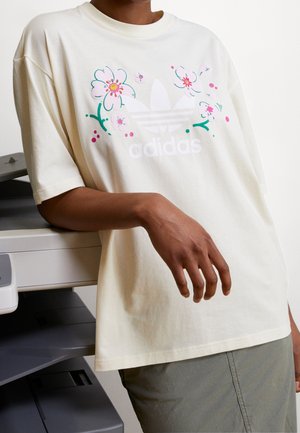 Henkilö nojaa tulostimeen, pukeutunut kermaiseen Adidas-t-paitaan, jossa on kukkakuvio logon ympärillä, ja oliivinvihreisiin housuihin.