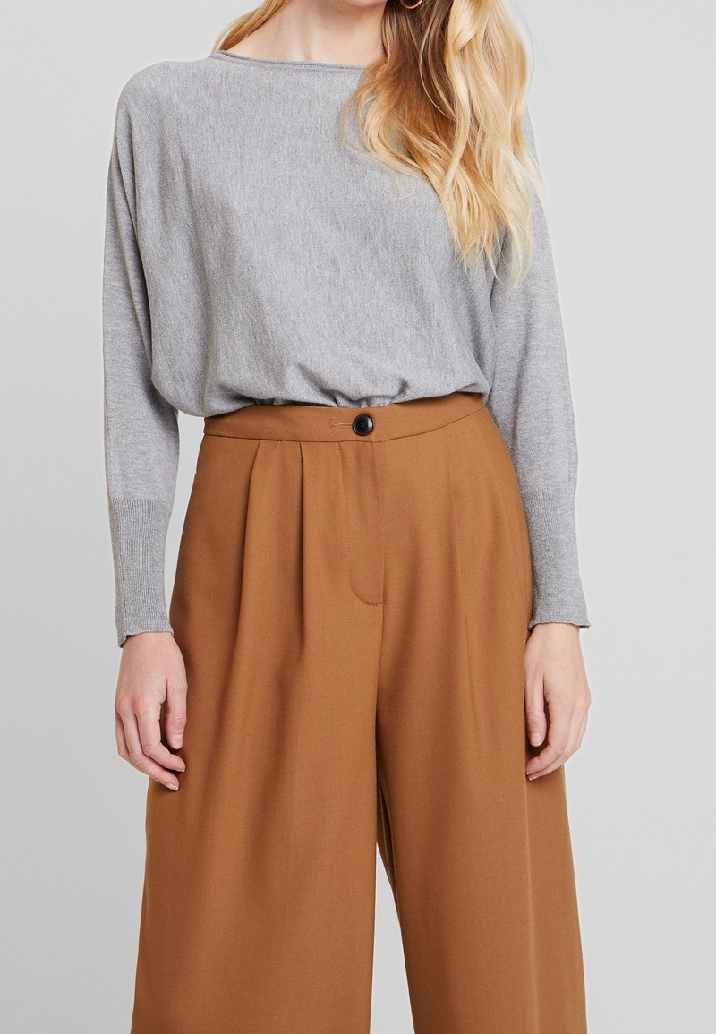 Femme portant un haut gris clair à manches longues rentré dans un pantalon marron taille haute à jambes larges avec un bouton noir.
