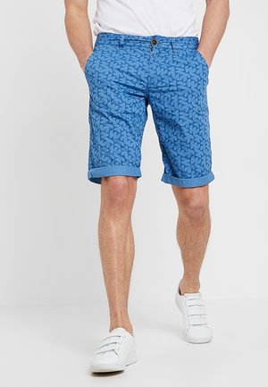 Man in blauwe geprinte shorts met opgerolde boorden, witte sneakers en een wit t-shirt, staand met handen in de zakken.