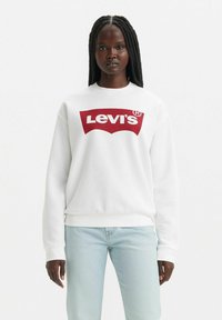 Vit sweatshirt med röd Levi's-logotyp, rund halsringning, ribbade ärmslut och nederkant. Bärs med ljusblå jeans. Enkelt och avslappnat design.
