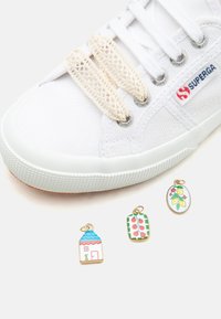 Λευκό αθλητικό παπούτσι Superga με μπεζ υφασμάτινους σιλικόνης κλειδαριές κορδονιών χωρίς δέσιμο και τρία μικρά πολύχρωμα σμάλτα γούρια από κάτω.