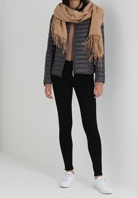 Giacca a vento grigia scura con cuciture orizzontali, abbinata a una sciarpa frangiata color sabbia, jeans skinny neri e sneakers bianche.