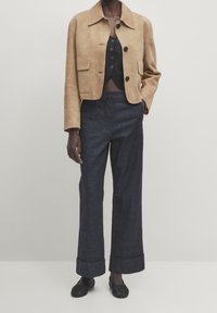 Veste en daim couleur fauve avec col, poches avant, et jean large bleu foncé avec revers roulés. Le haut est un crop top noir à boutons.