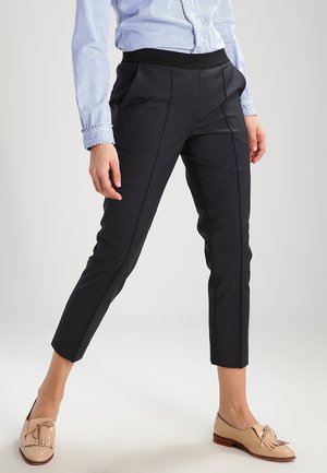 Kvinde iført sorte skræddersyede ankellange bukser, lyseblå stribet skjorte med knapper og beige loafers med kvaster, stående med benene spredt.