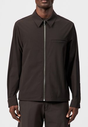 Mężczyzna ubrany w ciemnobrązową koszulę z długim rękawem, z przednim zamkiem błyskawicznym i małą kieszenią na piersi, w połączeniu z pasującymi ciemnobrązowymi spodniami.