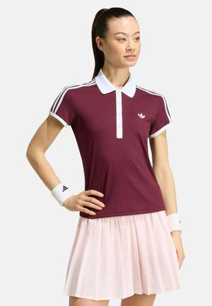Vrouw draagt een bordeauxrode Adidas poloshirt met witte kraag en strepen, een lichtroze geplooide rok en witte polsbandjes.