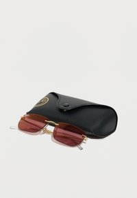 Γυαλιά ηλίου με χρυσή διπλή σκελετό και κόκκινους φακούς που ακουμπούν δίπλα σε μια κλειστή μαύρη υφασμάτινη θήκη με κουμπί snap και χρυσά γράμματα λογότυπου.