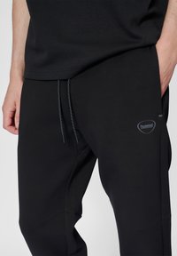 Schwarze Sporthose aus weichem Stoff, mit einem Kordelzugbund, seitlichen Taschen und einem kleinen Logo am Unterschenkel.
