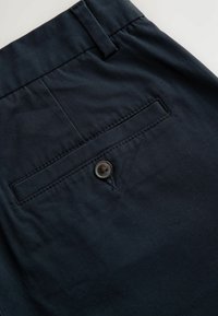 Pantalons bleu marine foncé avec une taille plate, des poches latérales et une poche arrière avec un bouton unique. Tissu lisse avec un léger éclat.