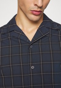 Camisa de cuadro marino de manga corta con cuello en pico, detalle sutil de botones y líneas blancas en contraste. Fabricada en mezcla de algodón, con textura suave.