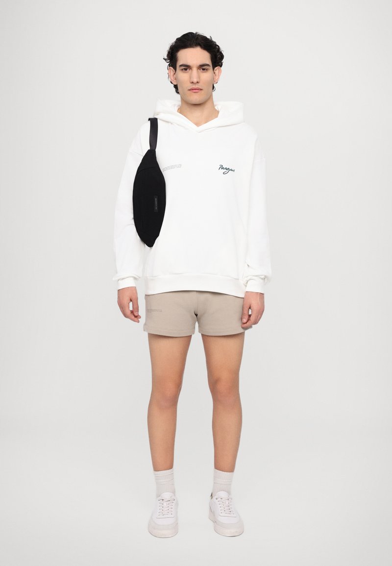 Bílá mikina s potiskem, béžové šortky a černá křížová taška. Bílé tenisky doplňují outfit. Jednoduchý, pohodlný streetwearový vzhled.
