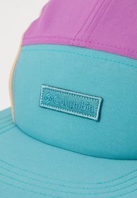 Căciulă colorată cu un panou frontal violet, vizieră teal și accente bej, având un logo Columbia embosat pe partea din față.