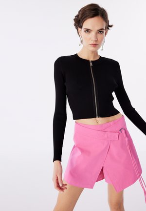 Pull cropped côtelé noir à manches longues avec fermeture éclair argentée ; associé à un short en tissu rose clair avec un nœud sur le côté et des accents de couleur.
