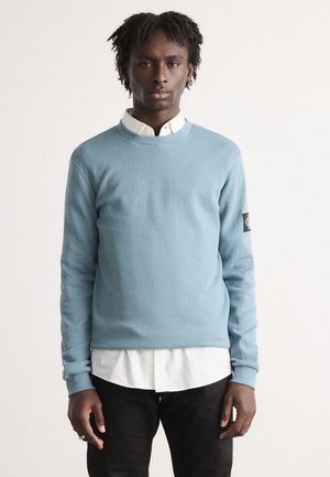 Ljusblå texturerad sweatshirt med rund hals, svart logotypapplikation på vänster ärm, bärs över en vit skjorta med krage och svarta byxor.