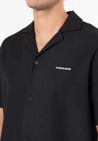 Maglietta nera a maniche corte con texture a coste, dotata di colletto con scollo a V, bottoni neri e logo bianco "PEGADOR" sul lato sinistro.
