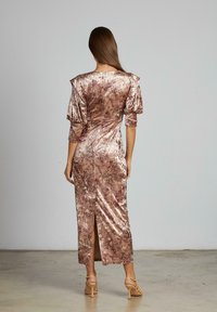 Donna con lunghi capelli castani che indossa un abito in velluto mauve aderente e lucido, con maniche a sbuffo e uno spacco sulla schiena, in piedi su un pavimento di cemento.