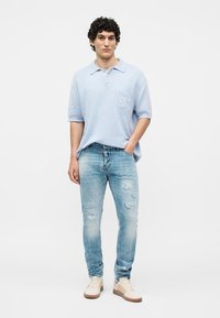 Homme debout portant un polo bleu clair, un jean bleu clair usé et des baskets beiges, une main dans la poche.