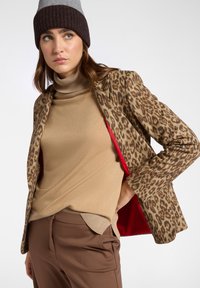 Giacca con stampa leopardata e interno rosso, dolcevita beige e pantaloni marroni. La giacca presenta spalle strutturate e una vestibilità su misura.