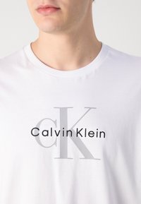 Primer plano de una persona que lleva una camiseta blanca con un gran "cK" gris y un logo de texto negro de "Calvin Klein" en el frente.