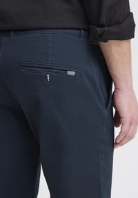 Muž ve tmavě modrých kalhotách s knoflíkovou zadní kapsou a černou košilí s vyhrnutými rukávy.