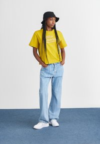 Gul kortermet t-skjorte med "New Balance"-logo, sammen med lys blå stripete jeans og hvite sneakers. Svart bøttehatt brukt.