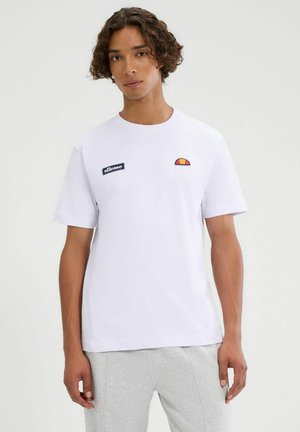 Junger Mann mit lockigem Haar, der ein weißes Ellesse-T-Shirt und hellgraue Hosen trägt, vor einem einfarbigen Hintergrund steht.