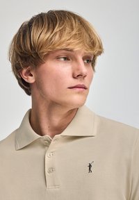 Polo beige con tejido texturizado, tres botones y un pequeño logo bordado de un golfista en la parte izquierda del pecho.