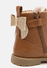 Bottine basse en cuir synthétique marron avec un accent de nœud doré pailleté, fermeture éclair latérale et semelle en caoutchouc texturée, présentant des détails à bords festonnés.