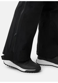 Zwarte waterdichte broek met ritsen aan de zijkanten, gecombineerd met zwart-witte wandelschoenen met gripzolen en vetersluiting.