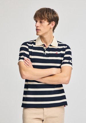 Polo a rayas en azul marino y crema con cuello clásico, mangas cortas y un pequeño logo rojo en el pecho. Ajuste casual y tela suave.