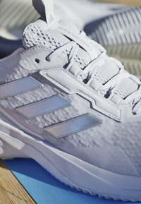 Weiße Sportschuhe mit einer strukturierten Oberseite, reflektierenden silbernen Akzenten und einem gestuften Design. Enthält Schnürsenkel und eine gepolsterte Sohle.